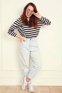 Genç güzel esmer kadın kız modeli yaz hipster giysiler ve güneş gözlüğü duvara yakın poz çıplak makyaj ile portresi. Delirmek ve dilini gösteren