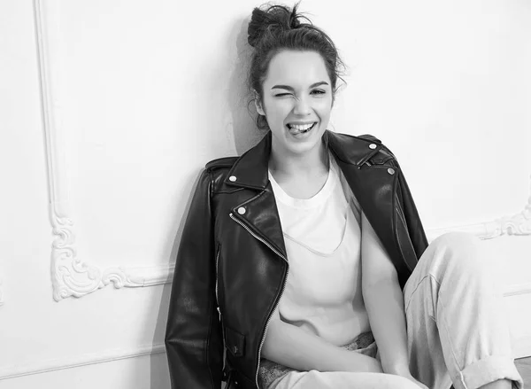 Portrait of young beautiful brunette woman girl model with nude makeup wearing summer hipster biker leather jacket clothes posing near wall. Sitting on the floor and winking