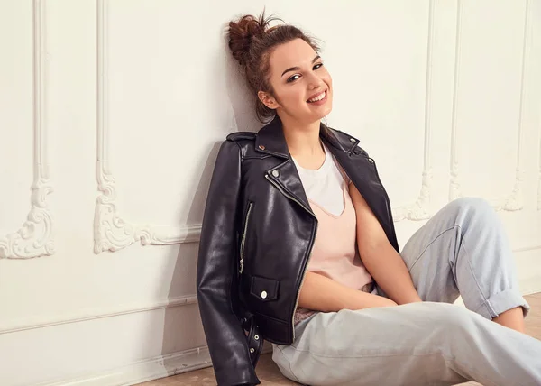 Portrait of young beautiful brunette woman girl model with nude makeup wearing summer hipster biker leather jacket clothes posing near wall. Sitting on the floor