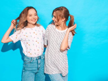 İki genç trendy sarışın hipster kızlarda gülümseyen güzel renkli tişört elbise yaz. Seksi kaygısız kadın mavi duvara yakın poz. Olumlu modelleri eğleniyor