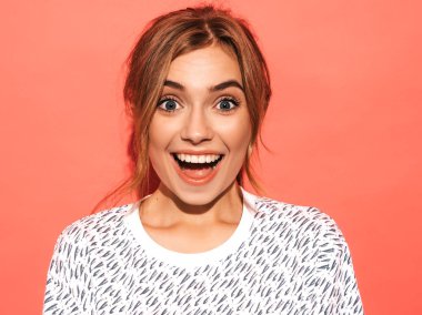 Genç ve güzel bir kadın kameraya bakıyor. Doğal makyajlı, günlük yazlık gömlekli moda bir kız. Gülümseyen olumlu bir kadın. Stüdyoda pembe duvarın yanında poz veren komik bir model. Şaşırmış ve şaşırmış.