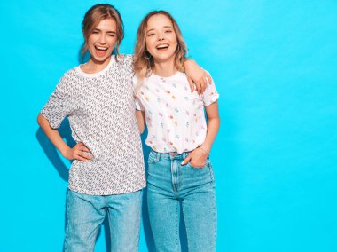 İki genç trendy sarışın hipster kızlarda gülümseyen güzel renkli tişört elbise yaz. Seksi kaygısız kadın mavi duvara yakın poz. Olumlu modelleri eğleniyor