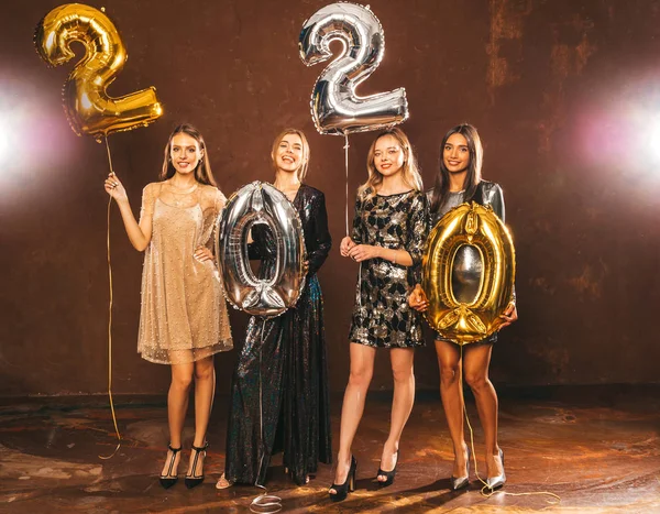 Beautiful Women Celebrating New Year. Happy Gorgeous Girls In Stylish Sexy Party Dresses Holding Gold and Silver 2020 Balloons, Having Fun At New Year\'s Eve Party. Holiday Celebration.Charming Models