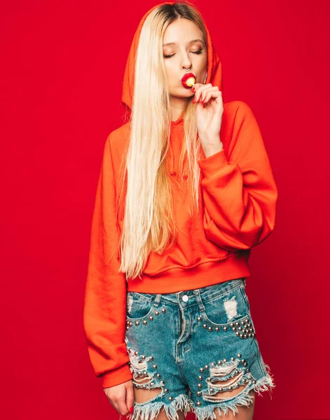 Portrait of young beautiful hipster bad girl in trendy red summer hoodie and earring in her nose.Sexy carefree smiling blond woman posing in studio.Positive model licking round sugar candy