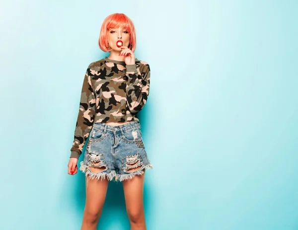 Portrait of young beautiful hipster bad girl in trendy jeans shorts and earring in her nose.Sexy carefree smiling woman in pink wig posing in studio.Positive model licking round sugar candy