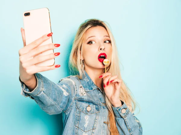 Young beautiful hipster bad girl in trendy jeans summer clothes and earring in her nose.Sexy carefree smiling blond woman posing in studio.Positive model licking round sugar candy.Takes selfie photo