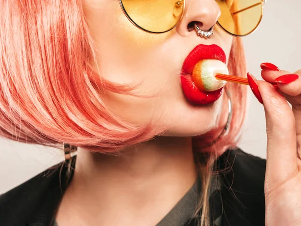Closeup lips of young beautiful hipster bad girl in trendy black leather jacket and earring in her nose.Sexy carefree smiling woman posing in studio in pink wig.Positive model licking round candy