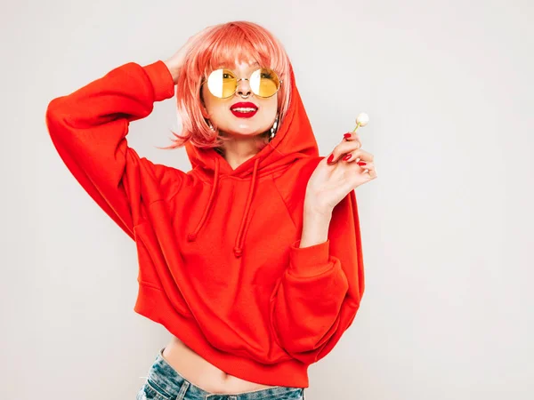 Portrait of young beautiful hipster bad girl in trendy red summer hoodie and earring in her nose.Sexy carefree smiling blond woman posing in studio in wig.Positive model licking round sugar candy