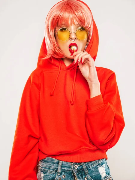 Portrait of young beautiful hipster bad girl in trendy red summer hoodie and earring in her nose.Sexy carefree smiling blond woman posing in studio in wig.Positive model licking round sugar candy