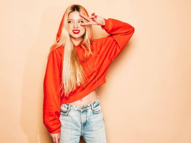 Portrait of young beautiful hipster bad girl in trendy red summer hoodie and earring in her nose.Sexy carefree smiling blond woman posing in studio.Positive model shows peace sign