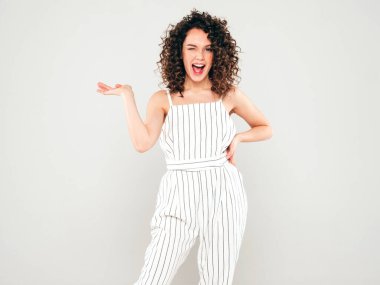 Portrait of beautiful smiling model with afro curls hairstyle dressed in summer hipster clothes.Sexy carefree girl posing in studio on gray background.Trendy woman shows something on her hand