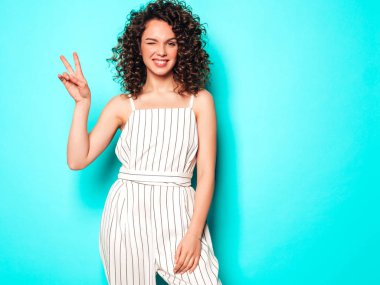 Kıvırcık saçlı güzel bir modelin portresi yaz hippi kıyafetleri içinde. Mavi duvarın yanındaki stüdyoda poz veren kaygısız kız. Moda, komik ve pozitif kadın barış işareti gösteriyor.