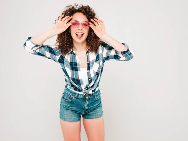 Portrait of beautiful smiling model with afro curls hairstyle dressed in summer hipster clothes.Sexy carefree girl posing in studio on gray background.Trendy woman shows tongue