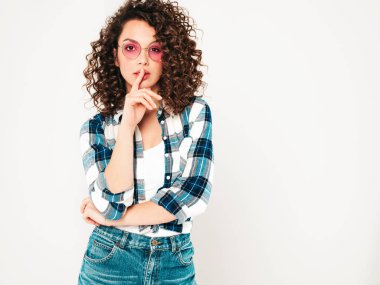 Portrait of beautiful smiling model with afro curls hairstyle dressed in summer hipster clothes.Sexy carefree girl posing in studio on gray background.Showes finger hush silence sign, gesture