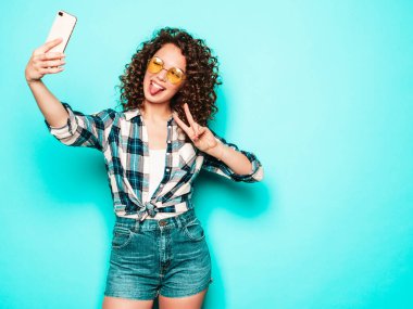 Portrait of beautiful smiling model with afro curls hairstyle dressed in summer hipster clothes.Sexy carefree girl posing in studio on gray background.Trendy funny woman takes selfie photo