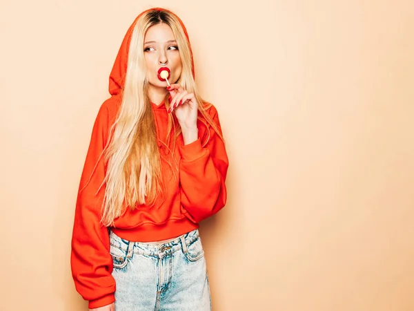 Portrait of young beautiful hipster bad girl in trendy red summer hoodie and earring in her nose.Sexy carefree smiling blond woman posing in studio.Positive model licking round sugar candy