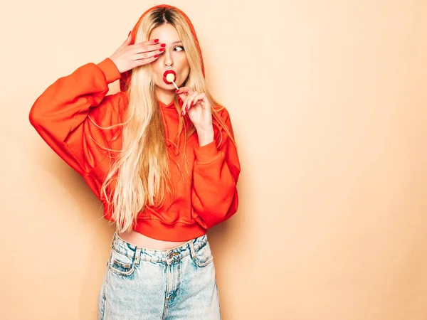 Portrait of young beautiful hipster bad girl in trendy red summer hoodie and earring in her nose.Sexy carefree smiling blond woman posing in studio.Positive model licking round sugar candy