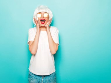 Young sexy smiling hipster girl in white wig and red lips.Beautiful trendy woman in summer clothes.Carefree model posing near blue wall in studio.Positive female going crazy in round sunglasses