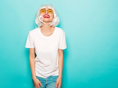 Young sexy smiling hipster girl in white wig and red lips.Beautiful trendy woman in summer clothes.Carefree model posing near blue wall in studio.Positive female going crazy in round sunglasses