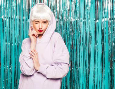 Young sexy smiling hipster girl in white wig and red lips.Beautiful woman in summer clothes.Carefree model posing on blue silver shiny tinsel background in studio.Positive female makes duck face