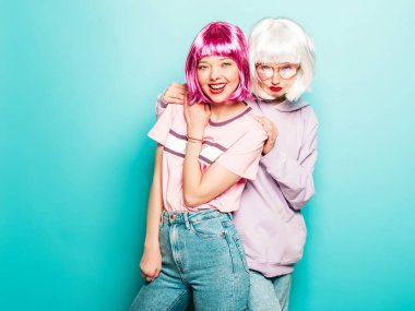 Two young sexy smiling hipster girls in wigs and red lips.Beautiful trendy women in summer clothes.Carefree models posing near blue wall in studio.Positive female going crazy and hugging