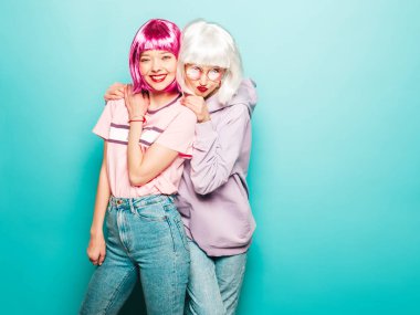 Two young sexy smiling hipster girls in wigs and red lips.Beautiful trendy women in summer clothes.Carefree models posing near blue wall in studio.Positive female going crazy and hugging