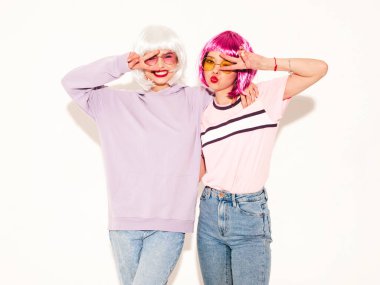 Two young sexy smiling hipster girls in wigs and red lips.Beautiful trendy women in summer clothes.Carefree models posing near white wall in studio.Positive female going crazy and shows peace sign