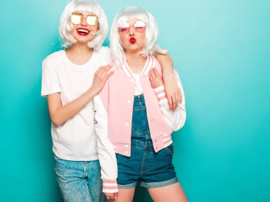 Two young sexy smiling hipster girls in wigs and red lips.Beautiful trendy women in summer clothes.Carefree models posing near blue wall in studio.Positive female going crazy and hugging