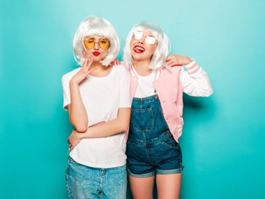 Two young sexy smiling hipster girls in wigs and red lips.Beautiful trendy women in summer clothes.Carefree models posing near blue wall in studio.Positive female going crazy and hugging