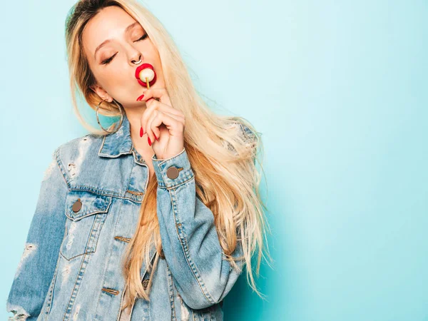 Portrait of young beautiful hipster bad girl in trendy jeans summer clothes and earring in her nose.Sexy carefree smiling blond woman posing in studio.Positive model licking round sugar candy