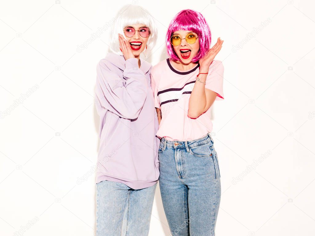 Two young sexy smiling hipster girls in wigs and red lips.Beautiful trendy women in summer clothes.Carefree models posing near white wall in studio.Positive female shocked and surprised