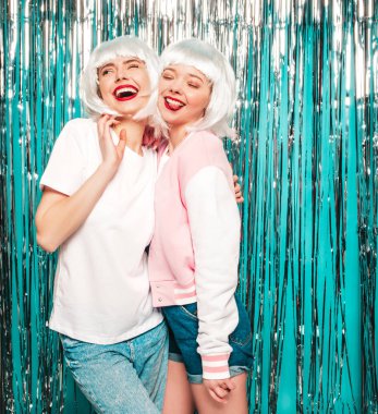 Two young sexy smiling hipster girls in white wigs and red lips.Beautiful trendy women in summer clothes.Carefree models posing on blue silver shiny tinsel background in studio.Positive female