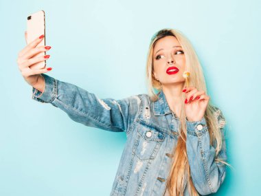 Genç, güzel, hippi kötü kız portresi moda kotlar içinde, yaz kıyafetleri içinde ve burnunda küpe. Kaygısız, güler yüzlü, sarışın kadın selfii alıyor. Şeker şekeri yuvarlak yalayan pozitif model.