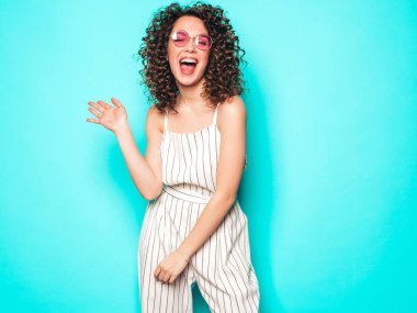 Kıvırcık saçlı, güzel bir mankenin portresi yaz hippi kıyafetleri giymiş, mavi duvarın yanında poz veren kaygısız bir kız. Güneş gözlüklü, modaya uygun ve pozitif bir kadın.