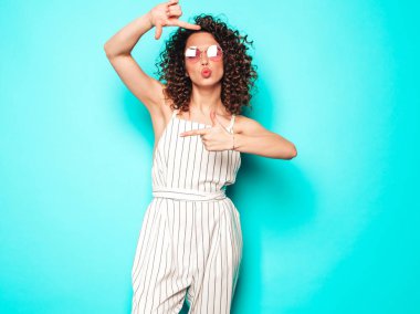 Portrait of beautiful smiling model with afro curls hairstyle dressed in summer hipster clothes.Sexy carefree girl posing near blue wall.Trendy woman makes photo frame by hands