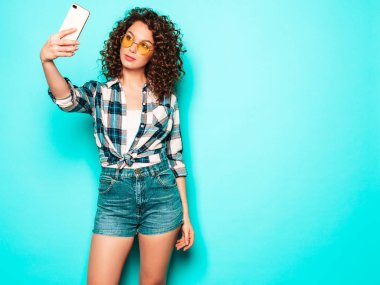 Portrait of beautiful smiling model with afro curls hairstyle dressed in summer hipster clothes.Sexy carefree girl posing in studio on gray background.Trendy funny woman takes selfie photo