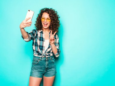 Portrait of beautiful smiling model with afro curls hairstyle dressed in summer hipster clothes.Sexy carefree girl posing in studio on gray background.Trendy funny woman takes selfie photo