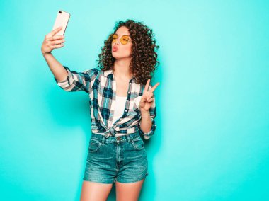 Portrait of beautiful smiling model with afro curls hairstyle dressed in summer hipster clothes.Sexy carefree girl posing in studio on gray background.Trendy funny woman takes selfie photo