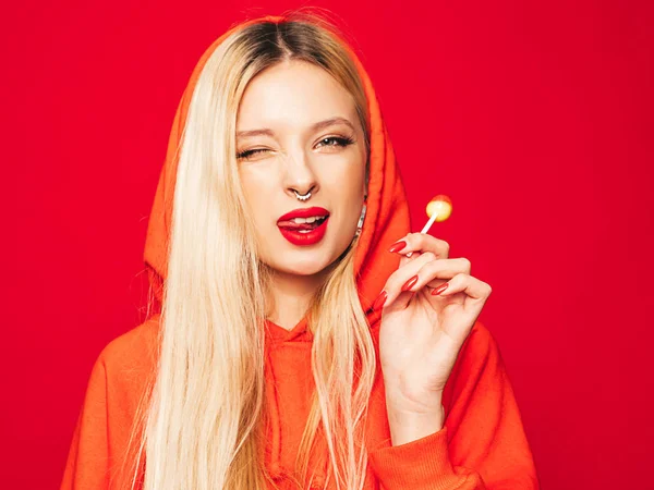 Portrait of young beautiful hipster bad girl in trendy red summer hoodie and earring in her nose.Sexy carefree smiling blond woman posing in studio.Positive model licking round sugar candy