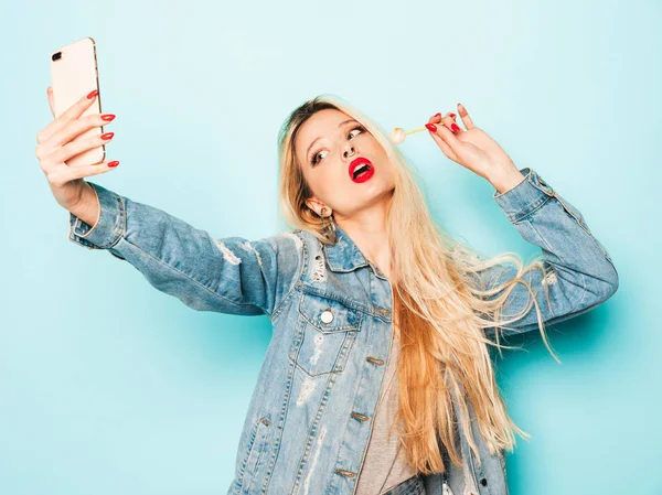 Portrait of young beautiful hipster bad girl in trendy jeans summer clothes and earring in her nose.Sexy carefree smiling blond woman takes selfie.Positive model licking round sugar candy