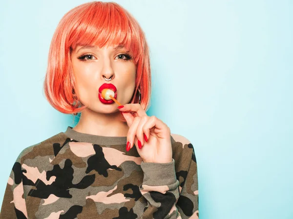 Portrait of young beautiful hipster bad girl in trendy jeans shorts and earring in her nose.Sexy carefree smiling woman in pink wig posing in studio.Positive model licking round sugar candy