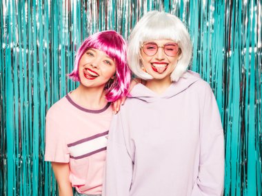 Two young sexy smiling hipster girls in white wigs and red lips.Beautiful trendy women in summer clothes.Carefree models posing on blue silver shiny tinsel background in studio.Positive female
