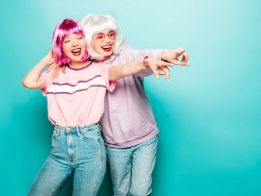 Peruklu ve kırmızı dudaklı iki genç seksi hippi kız. Yaz kıyafetleri içinde güzel moda kadınlar. Stüdyoda mavi duvarın yanında poz veren tasasız mankenler.
