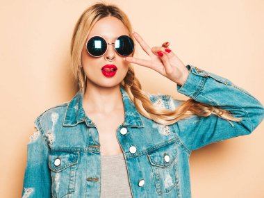 Genç, güzel, hippi kötü kız portresi moda kotlar içinde, yaz kıyafetleri ve burnunda küpe. Stüdyoda poz veren kaygısız, güler yüzlü, sarışın kadın. Güneş gözlükleriyle eğlenen pozitif model.