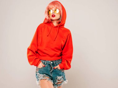 Güzel hippi kötü kız moda kırmızı yaz kapüşonlu ve burnunda küpeli. Pembe peruklu kaygısız kadın gri arka planda stüdyoda poz veriyor. Güneş gözlüklü seksi model.