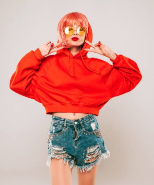 Güzel hippi kötü kız moda kırmızı yaz kapüşonlu ve burnunda küpeli. Pembe peruklu kaygısız kadın gri arka planda stüdyoda poz veriyor. Güneş gözlüklü seksi model.