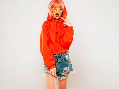 Güzel hippi kötü kız moda kırmızı yaz kapüşonlu ve burnunda küpeli. Pembe peruklu kaygısız kadın gri arka planda stüdyoda poz veriyor. Güneş gözlüklü seksi model.