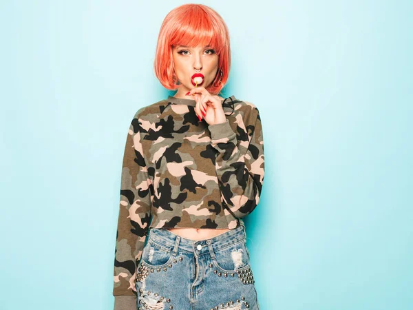 Portrait of young beautiful hipster bad girl in trendy jeans shorts and earring in her nose.Sexy carefree smiling woman in pink wig posing in studio.Positive model licking round sugar candy