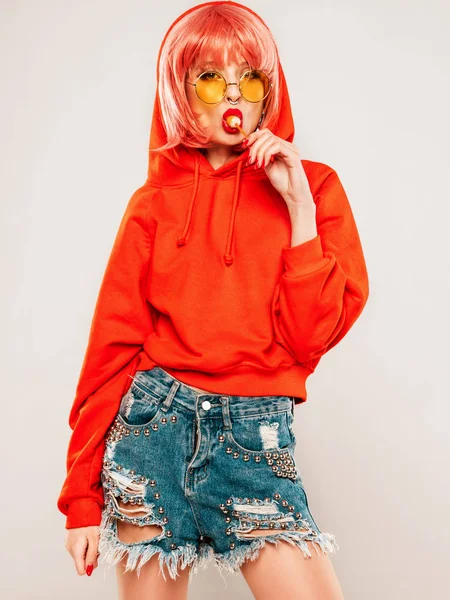 Young beautiful hipster bad girl in trendy red summer red hoodie and earring in her nose.Sexy carefree woman posing in studio on gray background in wig.Hot model licking round sugar candy