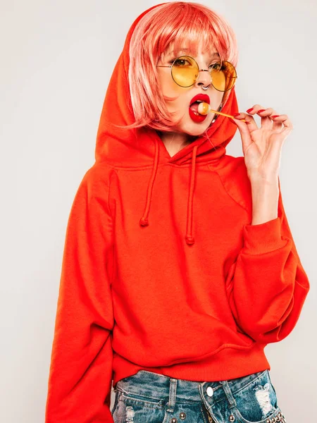 Young beautiful hipster bad girl in trendy red summer red hoodie and earring in her nose.Sexy carefree woman posing in studio on gray background in wig.Hot model licking round sugar candy